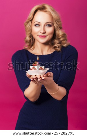 Beautiful blonde woman in luxury blue dress and curly hairstyle blowing candle on birthday cake. clear skin. pink background. focus on cake