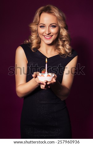 Beautiful blonde woman in luxury black dress and curly hairstyle blowing candle on birthday cake. clear skin. dark red background