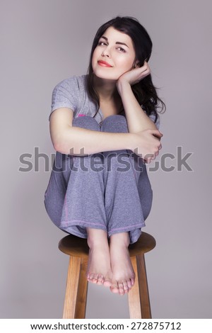 Brunette beautiful woman sitting on a chair home alone, happy. neutral grey background