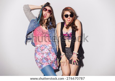Two beautiful brunette women (girls) teenagers spend time together having fun, make funny faces. Retro outfit: bright blue tights, jeans jacket, shorts in sequins, and colored strand of hair