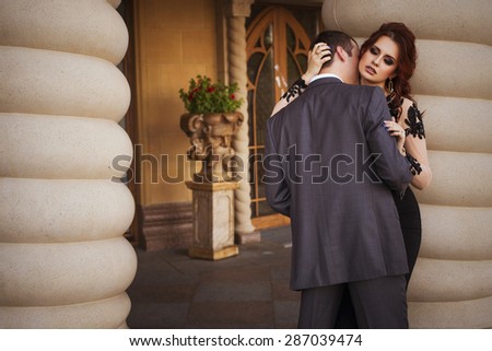 Beautiful sexy brunette luxury rich woman standing on a balcony in evening long black dress with a man in suit kissing her neck. Copy space