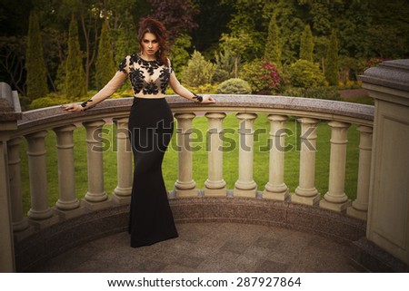 Beautiful sexy brunette luxury rich woman standing on a balcony near garden in evening long black dress. Copy space