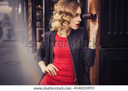 Beautiful brunette sexy spy agent (killer or police) woman in leather jacket and red luxury dress with a binocular in her hand stalking someone, to catch him in european city