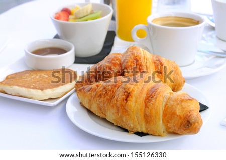 close-up of breakfast with croissants in white plate, coffee orange juice and fresh fruits in background