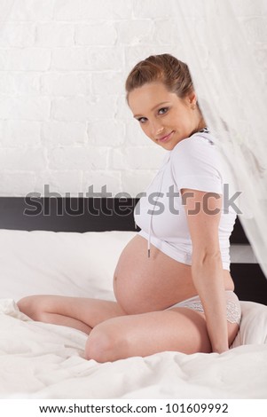 Pregnant woman sits on the house bed.