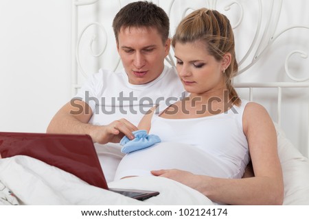 Happy pregnant family with notebook and children's bootees lies on the house bed.