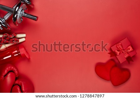 Dumbbells, boxing gloves, rope, bottle for water, gift box and two red hearts on a red background.Top view with copy space. Valentine\'s Day card. Fitness, sport and healthy lifestyle concept.