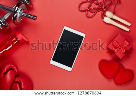Dumbbells, boxing gloves, rope, bottle for water, computer tablet, gift box and two hearts on red background.Top view with copy space. Valentine\'s Day card. Fitness, sport, healthy lifestyle concept.
