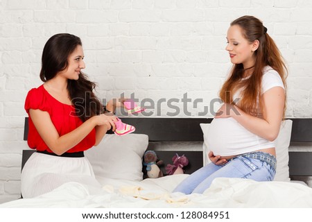 Two pregnant girlfriend  with children's clothes sit on the bed at home.