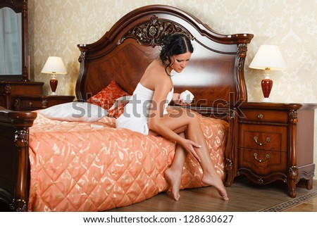 Young woman in towel with cosmetic cream sits on the house bed.  Concept of body care.