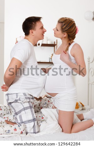 The happy pregnant woman with the husband sit on the house bed.