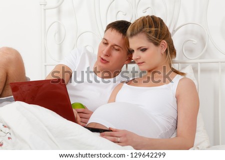 Happy pregnant family with notebook lies on the house bed.
