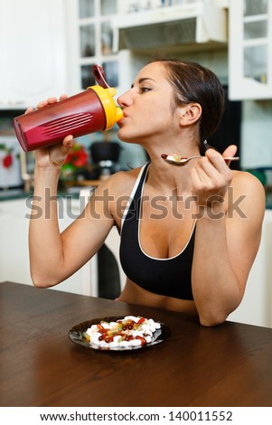 The sports young woman with a protein cocktail in a shaker and plate with nutritional supplements  sits in house kitchen. Sports nutrition.