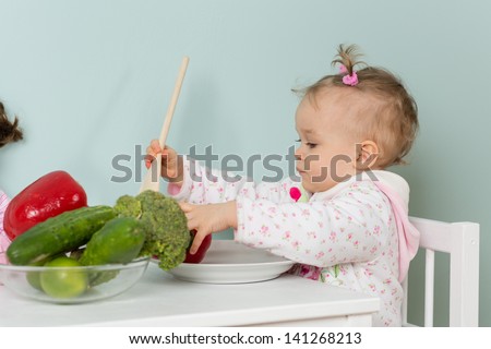 Sweet small child sits on a chair at the dining table with fresh vegetables in the home kitchen.