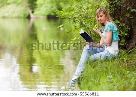 The beautiful young woman sits on a grass in park with the book and an apple.