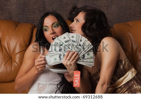 Young happy women with money sit on leather sofa at home. Shopping.