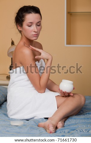The attractive young woman in towel with cosmetic cream sits on bed at home. Concept of body care.