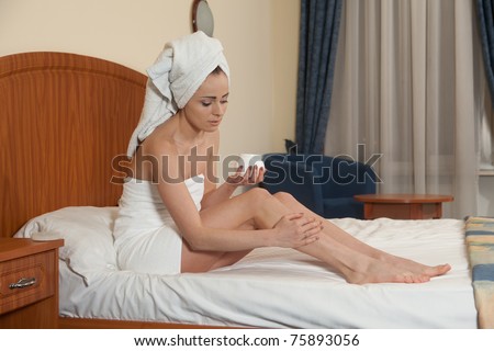 The attractive young woman in towel with cosmetic cream sits on bed at home. Concept of body care.
