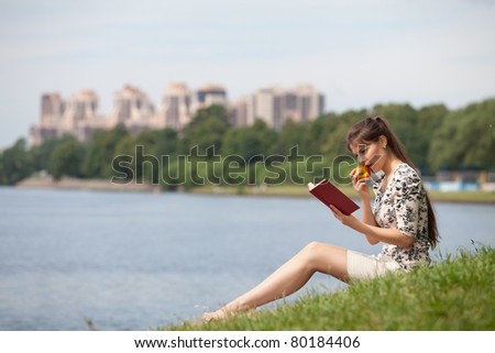 The beautiful young woman sits on a grass in park with the book and an apple.