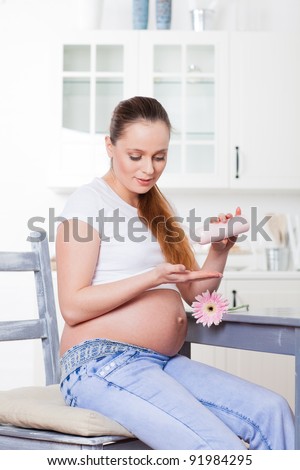 Young pregnant woman with cosmetic cream sits the  house kitchen. Concept of body care.