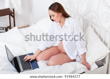 The beautiful young pregnant woman with laptop sits on the house bed.