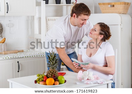 Pregnant family with money and baby\'s clothes sit in the house kitchen. Family budget.
