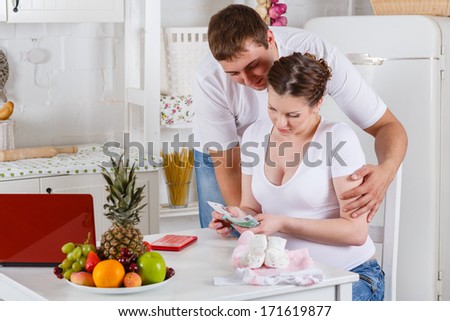 Pregnant family with money and baby\'s clothes sit in the house kitchen. Family budget.