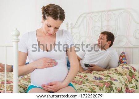 The pregnant woman becomes angry about the husband who lies on a bed and watches TV.