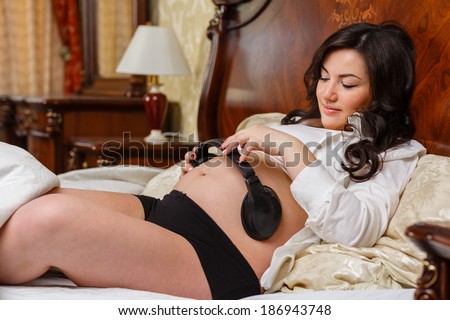 Pregnant woman with headphones lying on the bed and listening to music in the bedroom of the house.