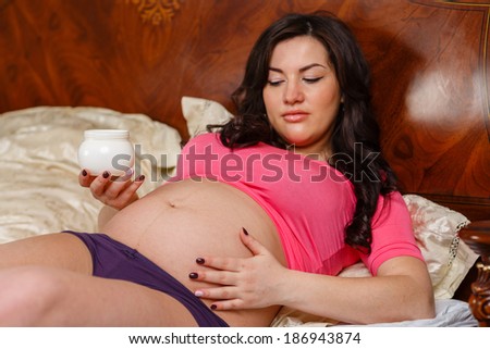 Young pregnant woman with cosmetic cream lies on a bed in the room. Selective focus on cream. Concept of body care.