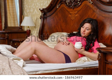 Young pregnant woman with cosmetic cream lies on a bed in the room. Concept of body care.