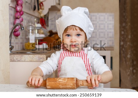 Little girl in apron and cap of the cook with rolling pin sits at the dining table in the kitchen in the house. Mother's helper. 2 year old.