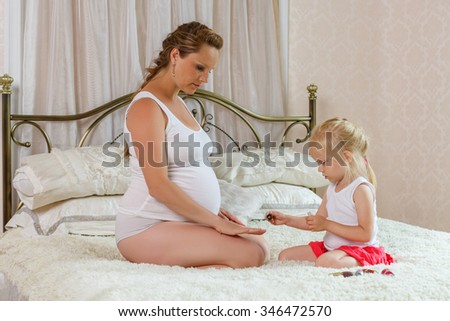 The little girl does a manicure to the pregnant mother in the bedroom of the house. Happy family.