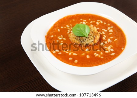 A picture of a white bowl full of fresh vegetable cream soup with pine nuts and piece of toast with basil on top