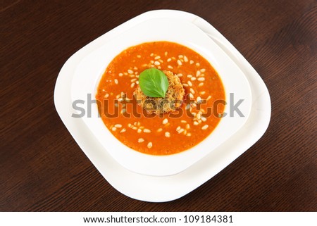 A picture of a white bowl full of fresh red pepper cream soup with pine nuts and piece of toast with basil on top