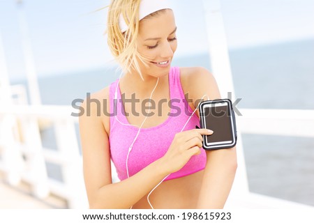Young fit woman in pink sports bra touching phone on the pier over sea