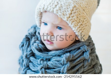 portrait of stylish pretty nice baby girl in wool snood and hat, tall boots and modern dress staying on white background and wooden floor watching aside
