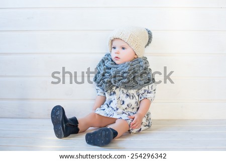 stylish pretty nice smiling baby girl in wool snood and hat, tall boots and modern dress sitting and posing on white background and wooden floor watching aside