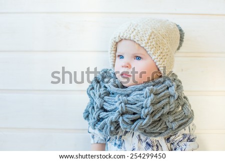 portrait of stylish pretty nice baby girl in wool snood and hat, tall boots and modern dress staying on white background and wooden floor watching aside