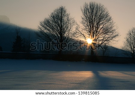 Sunset in mountains through trees along wall.  The sun sets behind a mountain ridge and beams shine through the trees, casting a shadow upon the snow covered ground.