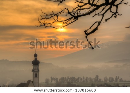 Sunrise on a foggy morning in the Alps.  Fog and mist in the mountains create both depth and color in the morning.  A tree in the foreground and church in the middle ground create a calming mood.