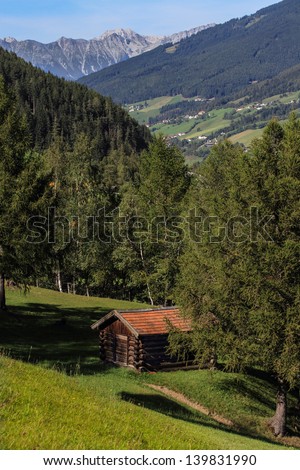 An isolated old hay shed up on the green timbered slopes of the Alps.  Small towns and big mountains can be seen in the distance.