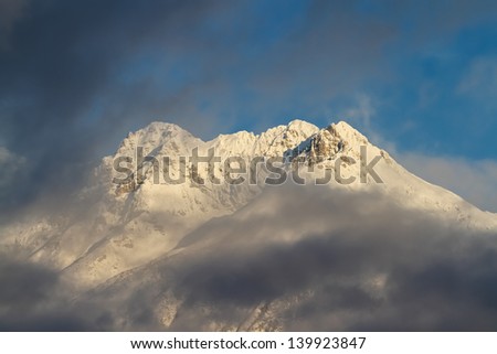 Early morning winter sun illuminates high mountain peak of the Alps above soft low clouds.