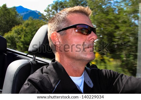 White male driving convertible in mountains with background blur from fast motion.