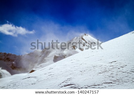 Winter Winds in the Alps.  Textured snow in the foreground with the wind blowing snow from a mountain summit into the blue sky.