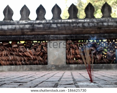 joss sticks smoke on the stone ground floor of a temple corridor terraces with temple wall with typical buddhism decorative stone leaf form ornament in the background in Vientiane, Laos