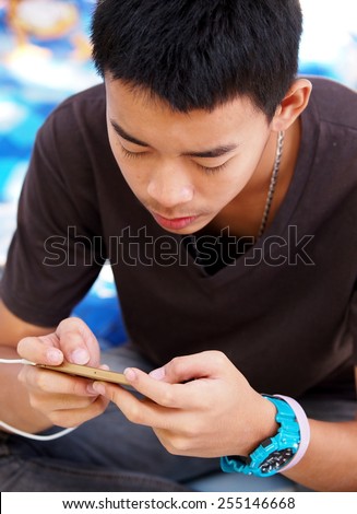 cute young asian boy with black short hairs wearing dark casual brown gray t-shirt blue sport watch sitting on the floor concentration on playing game and reading message on mobile handy smart phone