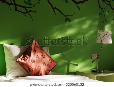 close up photo of a bed with white and red pillows in green painted wall bed room decorated with lamp and wall painting with natural light from glass windows showing garden shadows on the wall