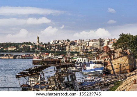 Belgrade downtown skyline with dominant St. Michael\'s Cathedral bell tower, contrasted to old, rusty, abandoned boats and barges at the ship junkyard on Sava river and Old Sava bridge in a distance.
