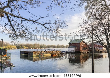 Early spring photograph shot at dusk of risen Sava river water level, with floating houses along its banks, leafless trees canopies, and blue skies with fluffy white clouds.